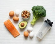 Kleurrijke flatlay van voedingsrijke producten: zalm, eieren, avocado, noten, broccoli en een eiwitshaker op wit gym-oppervlak.